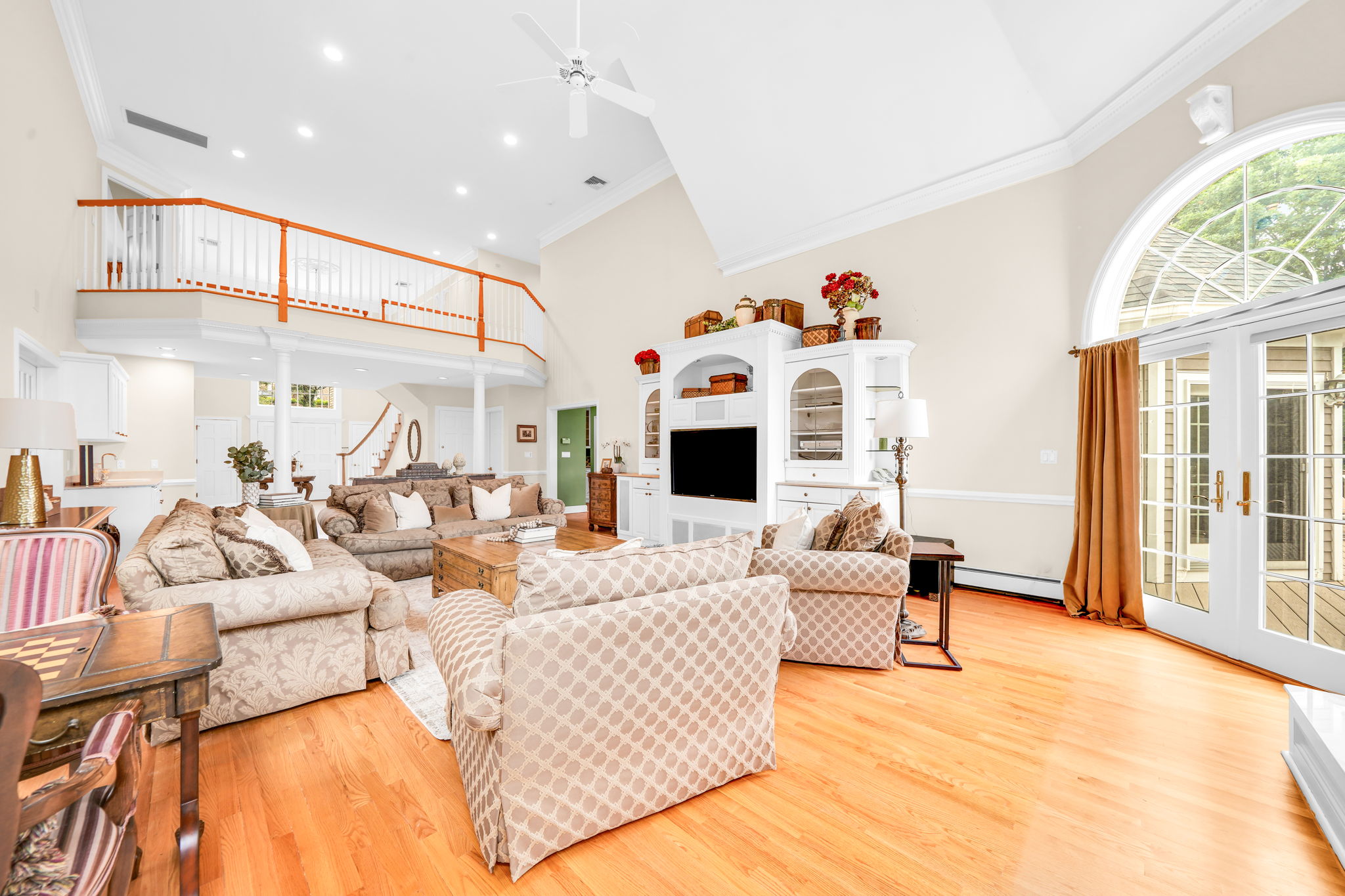 The great room itself is a showstopper: a soaring, two-story space beneath a dramatic vaulted ceiling with a graceful ceiling fan and elegant wall paneling throughout. A fireplace framed in detailed white millwork anchors the room, flanked by arched, palladian-style windows dressed in rich gold drapery, while double French doors open onto an expansive back deck and the flat, grassy backyard beyond. Rich hardwood floors and built-in cabinetry complete a space made for both grand entertaining and quiet evenings in.