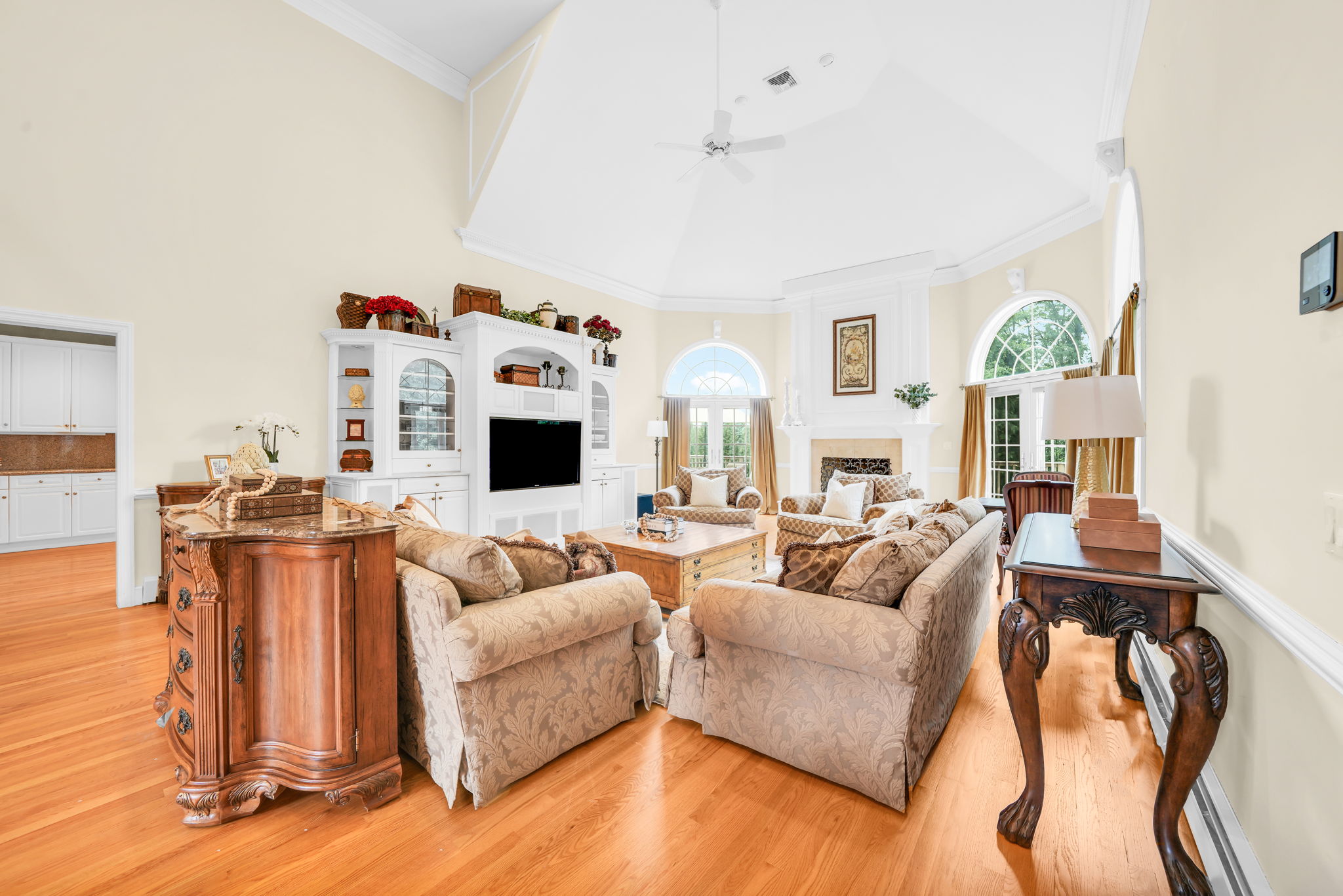 The great room itself is a showstopper: a soaring, two-story space beneath a dramatic vaulted ceiling with a graceful ceiling fan and elegant wall paneling throughout. A fireplace framed in detailed white millwork anchors the room, flanked by arched, palladian-style windows dressed in rich gold drapery, while double French doors open onto an expansive back deck and the flat, grassy backyard beyond. Rich hardwood floors and built-in cabinetry complete a space made for both grand entertaining and quiet evenings in.