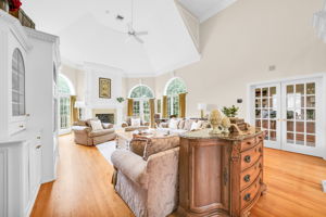 The great room itself is a showstopper: a soaring, two-story space beneath a dramatic vaulted ceiling with a graceful ceiling fan and elegant wall paneling throughout. A fireplace framed in detailed white millwork anchors the room, flanked by arched, palladian-style windows dressed in rich gold drapery, while double French doors open onto an expansive back deck and the flat, grassy backyard beyond. Rich hardwood floors and built-in cabinetry complete a space made for both grand entertaining and quiet evenings in.