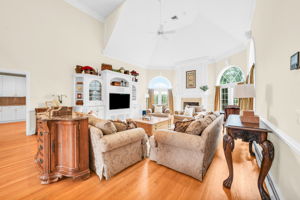 The great room itself is a showstopper: a soaring, two-story space beneath a dramatic vaulted ceiling with a graceful ceiling fan and elegant wall paneling throughout. A fireplace framed in detailed white millwork anchors the room, flanked by arched, palladian-style windows dressed in rich gold drapery, while double French doors open onto an expansive back deck and the flat, grassy backyard beyond. Rich hardwood floors and built-in cabinetry complete a space made for both grand entertaining and quiet evenings in.